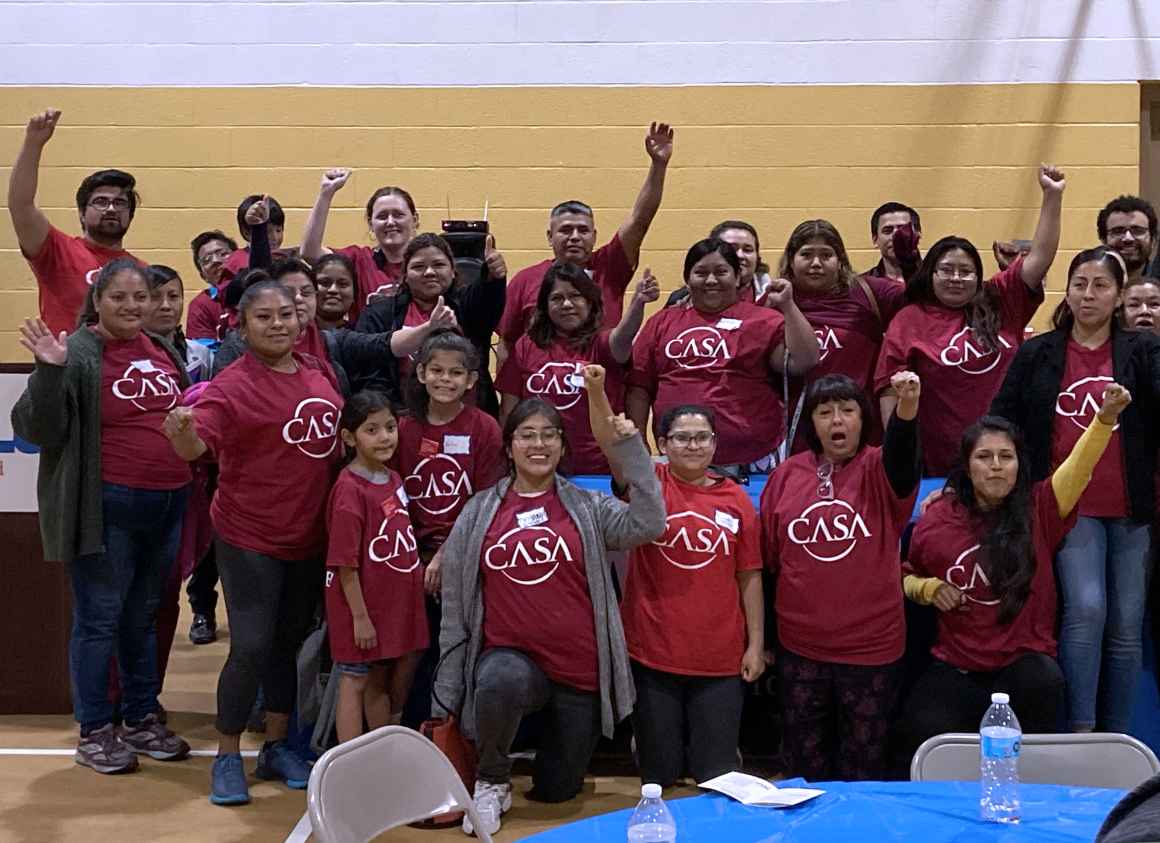 A group photo of CASA members wearing red CASA t-shirts. Several people have their first raised.
