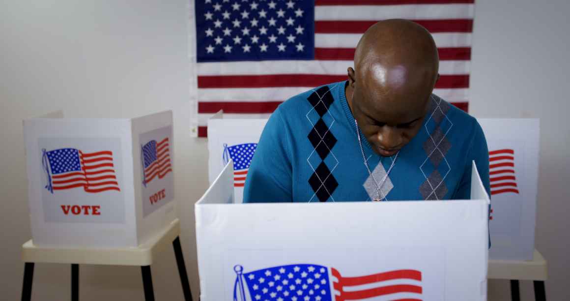 MS front view African American man in blue sweater and wearing a silver cross on a chain casting vote in booth at polling station. US flag on wall in background