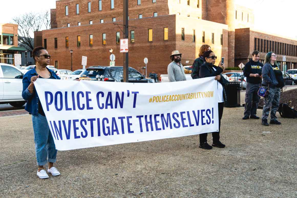 Two Black advocates hold up a banner that says, "Police Can't Investigate Themselves." They're standing outside at a rally.