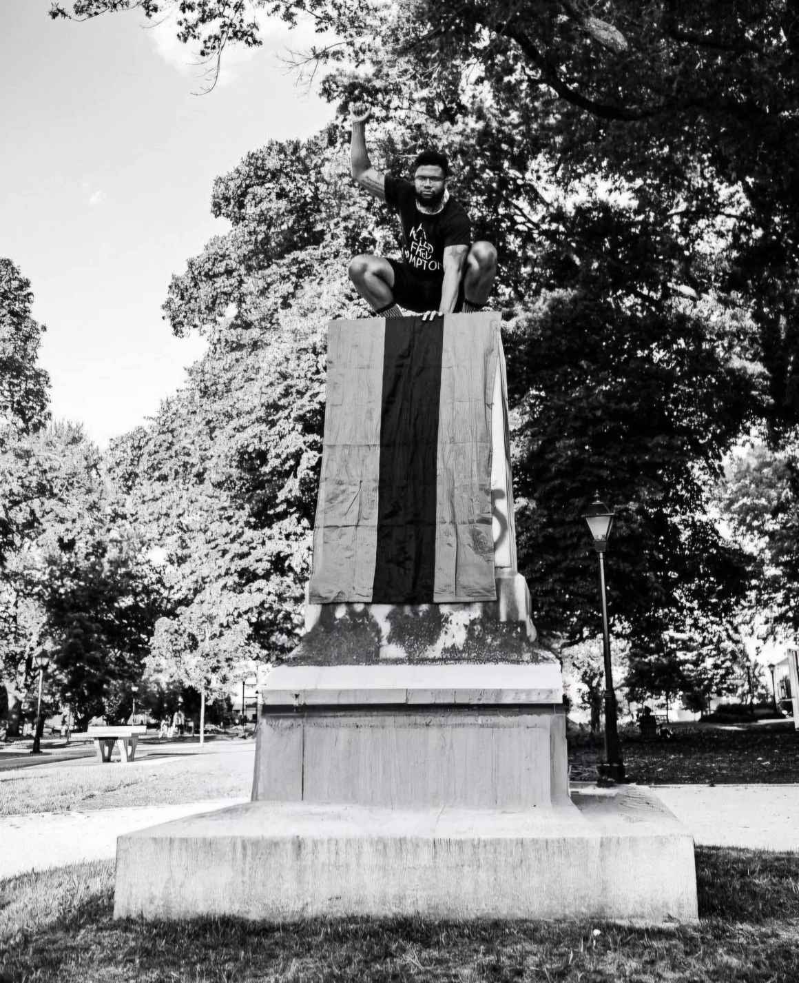 Black and white photo of Nehemiah Bester on top of a statue pedestal of a removed confederate statue. The Pan-African flag drapes over the pedestal.